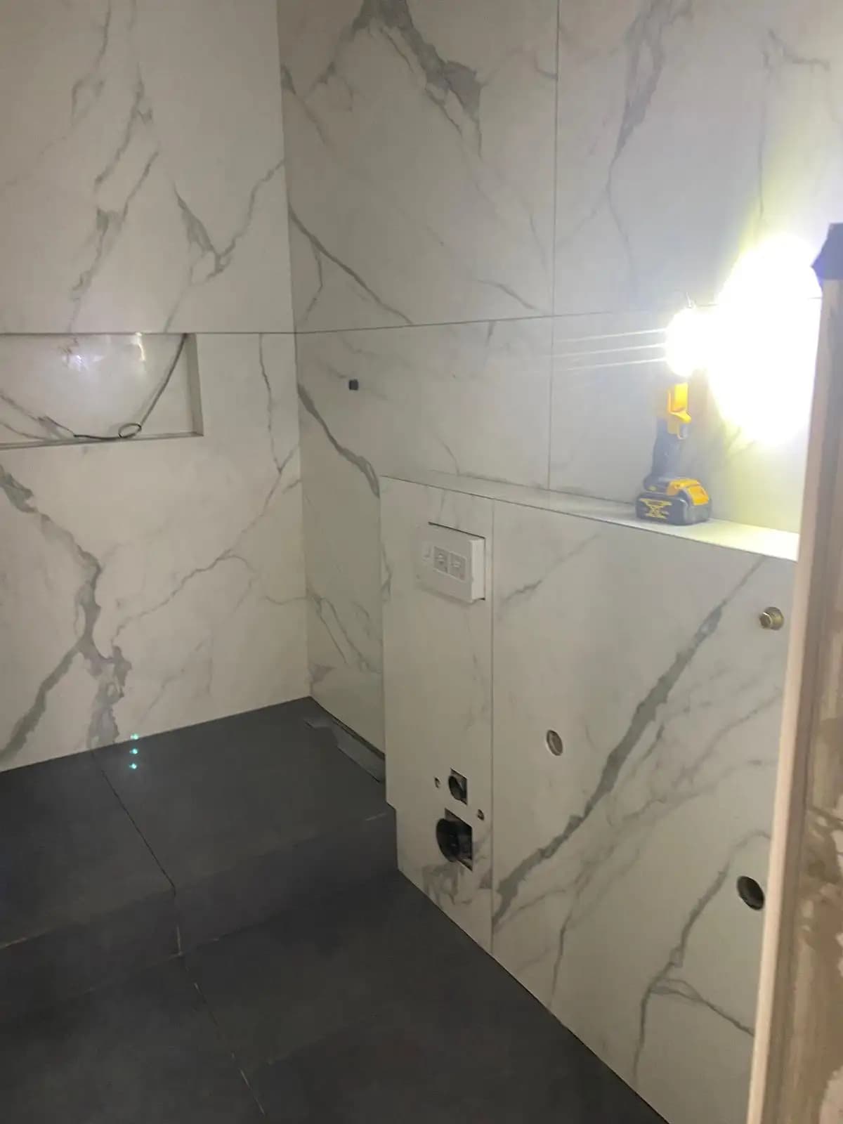 The image shows a bathroom undergoing renovation. A marble-tiled wall and flooring are visible, as well as a shower...