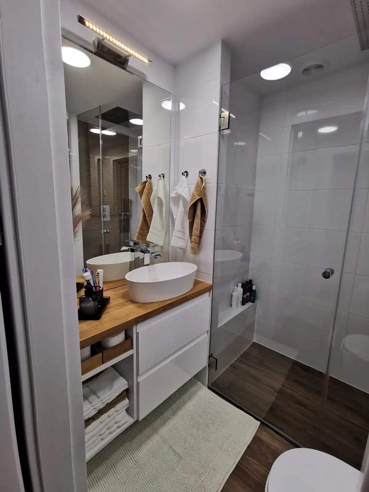 A small, modern bathroom with a white sink and countertop, a large rectangular mirror, a glass shower door, a tiled...