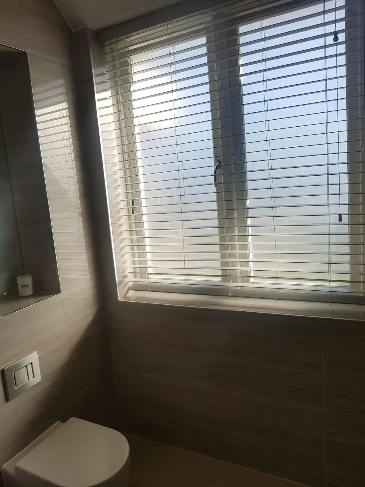 Small bathroom with white toilet and a window featuring white blinds and beige vertical blinds. The walls are tiled...