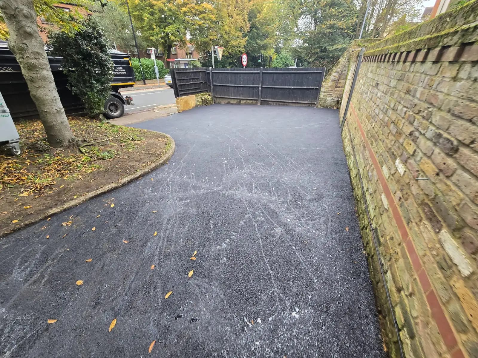 The image shows a view of an outdoor space with a paved area leading to a stone wall and driveway. There's a black...