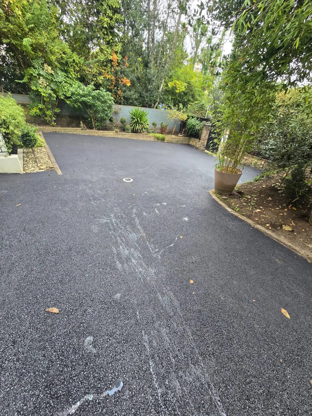 The image shows a driveway with gravel, surrounded by lush greenery and a few plants. A black asphalt surface leads up...