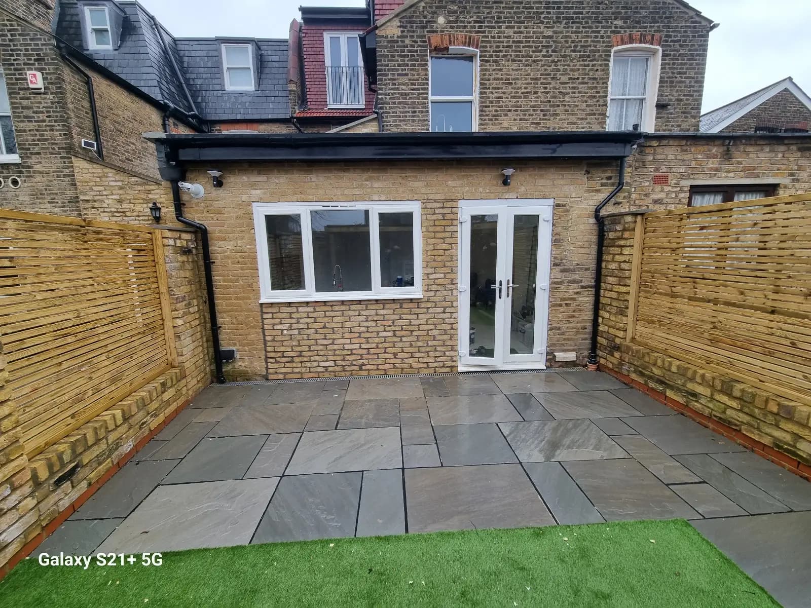 The image shows a backyard area with a brick patio and a wooden fence. There's an outdoor door on the right side of...