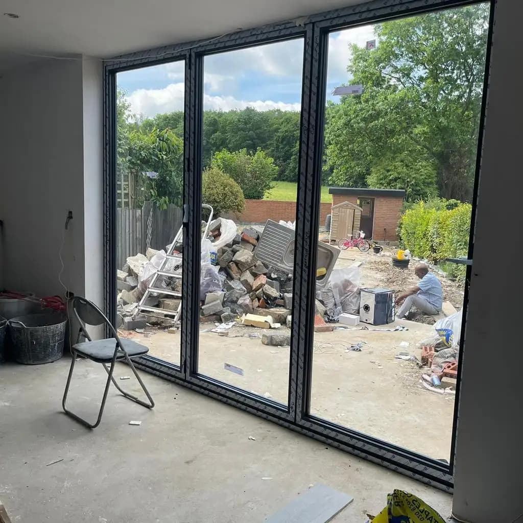 Sliding glass doors lead to a backyard under renovation with piles of construction materials and debris. A chair is...