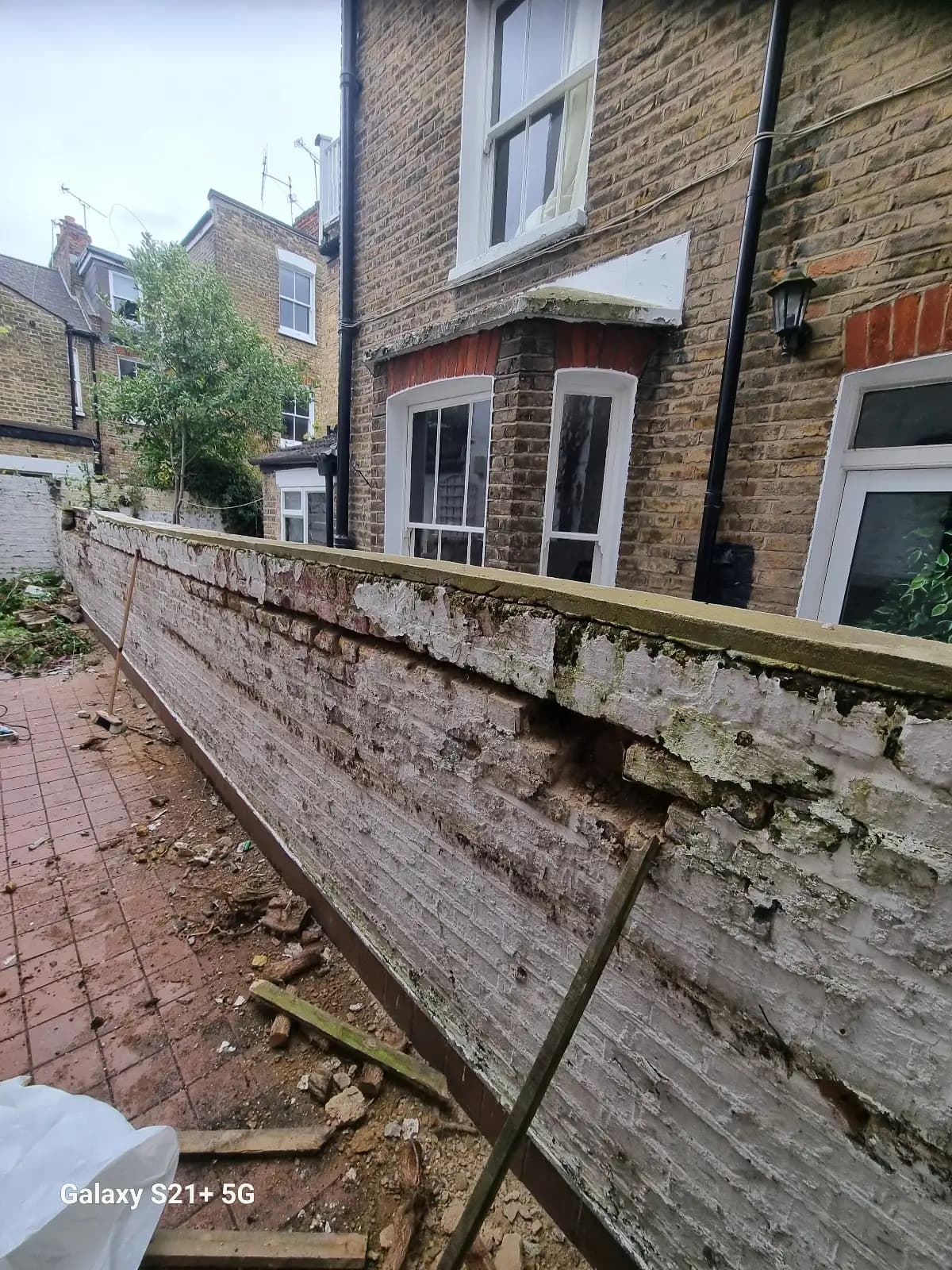 The image shows a house under renovation with visible wall damage and an old white fence. A brick wall appears to be...