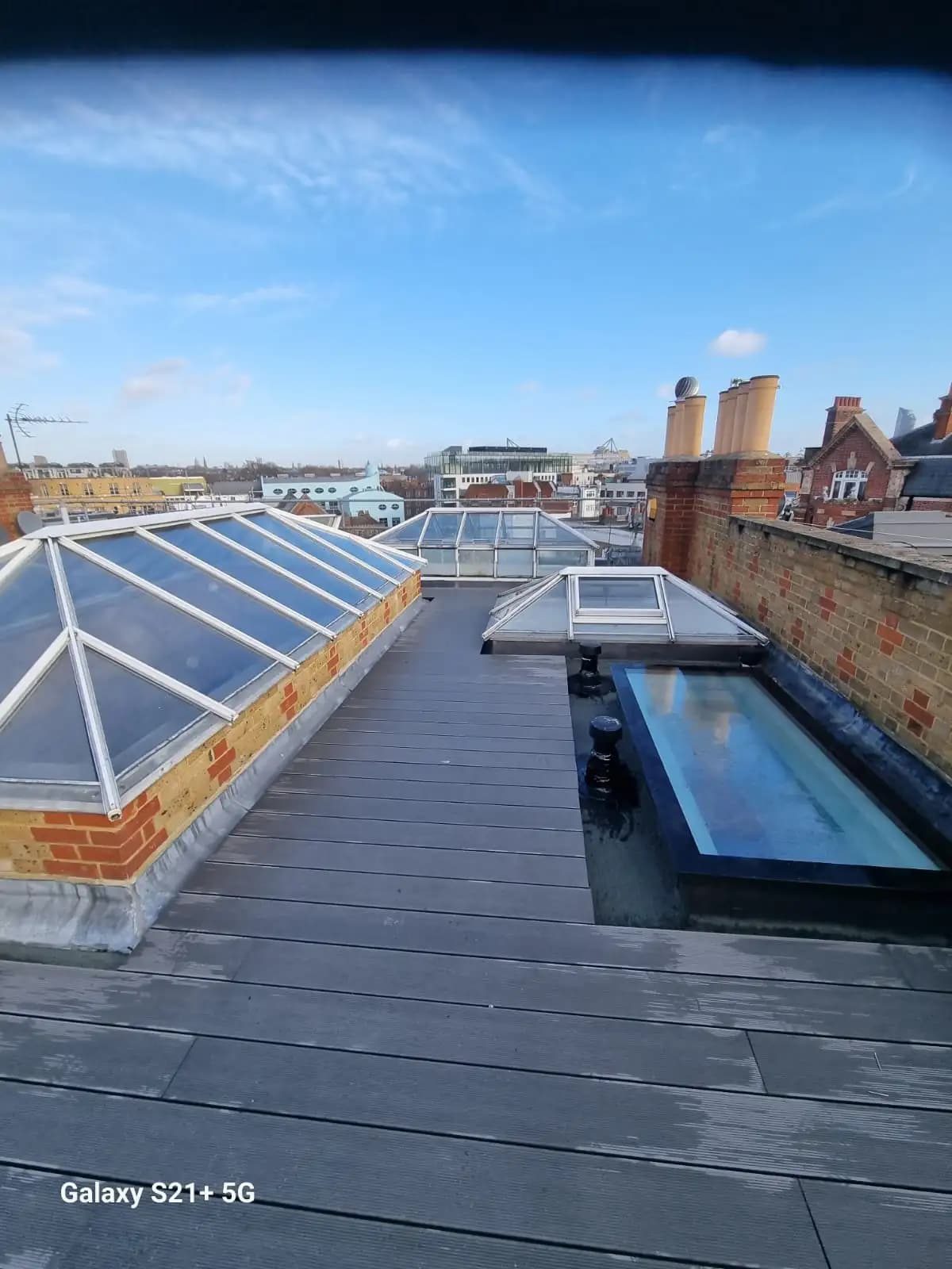 Alt Text: A sunny rooftop with a wooden deck and two skylights. The sky is partly cloudy, and there are chimneys in...