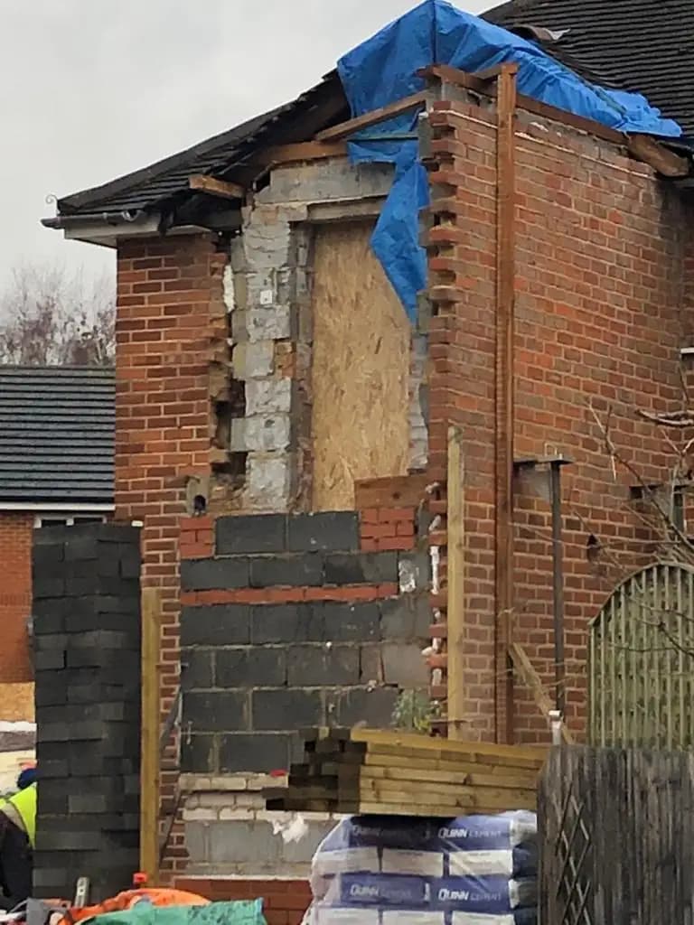 The image depicts a building under construction with a partial roof and exposed brickwork. A blue tarp covers an upper...