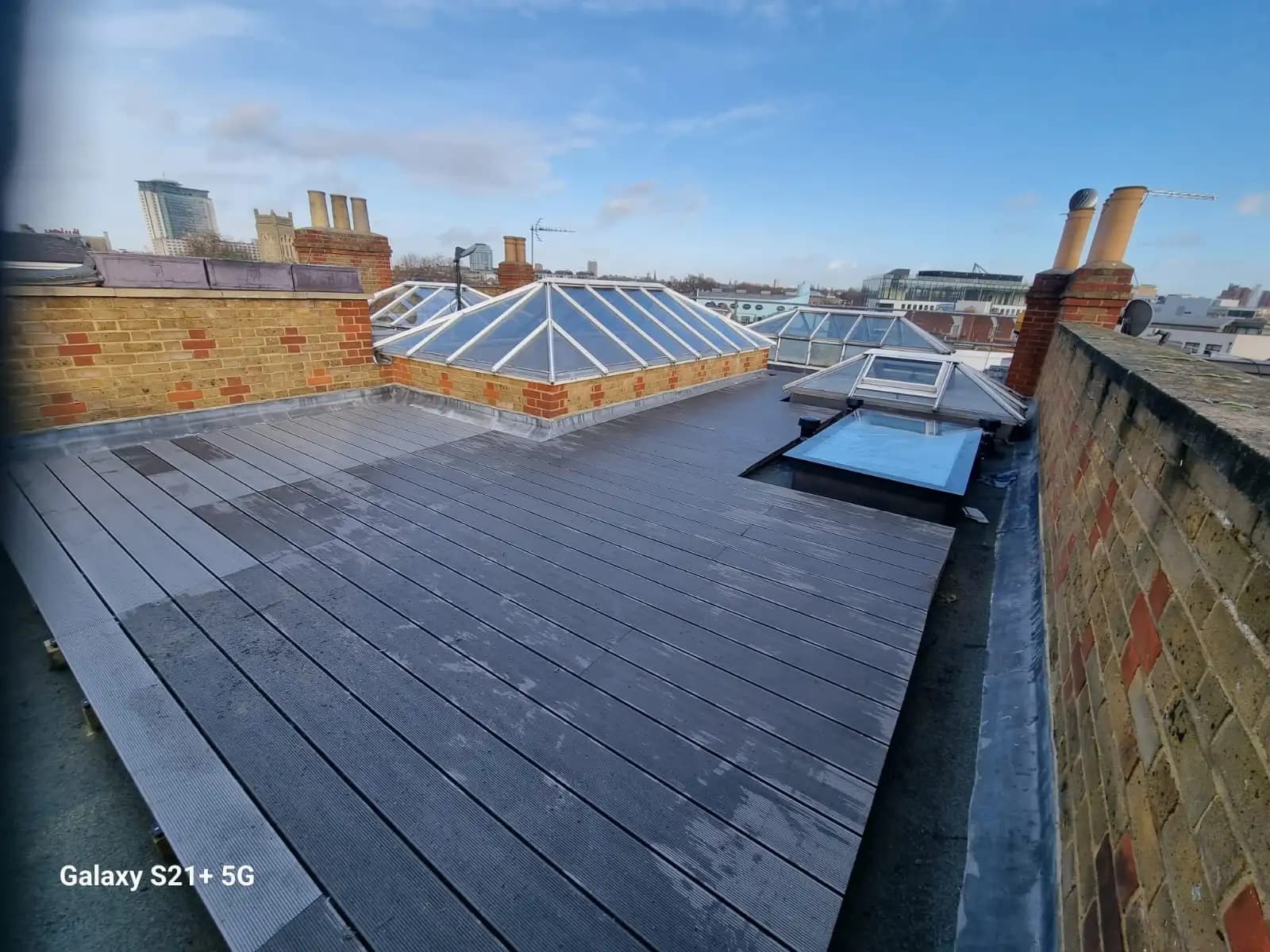 The image shows a rooftop with a wooden deck and a greenhouse-like structure in the background. There are also some...