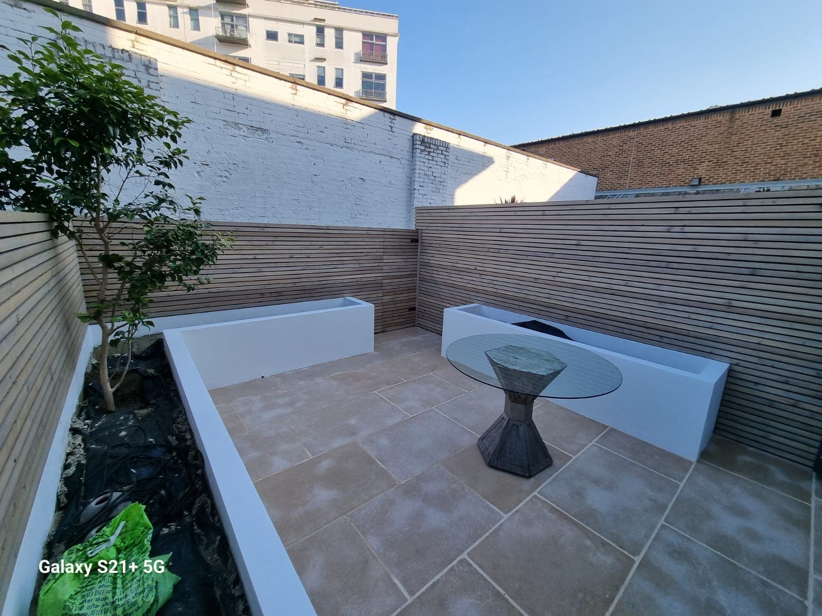 Outdoor area with patio furniture and a small table, featuring a deck with wooden flooring and a black circular ottoman. The patio is set against a building with white walls and has a garden area in the background.
2. Garden Construction. 